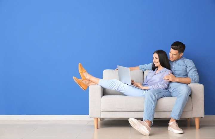 Un couple utilisant un PC portable sur un canapé, devant un mur bleu simple